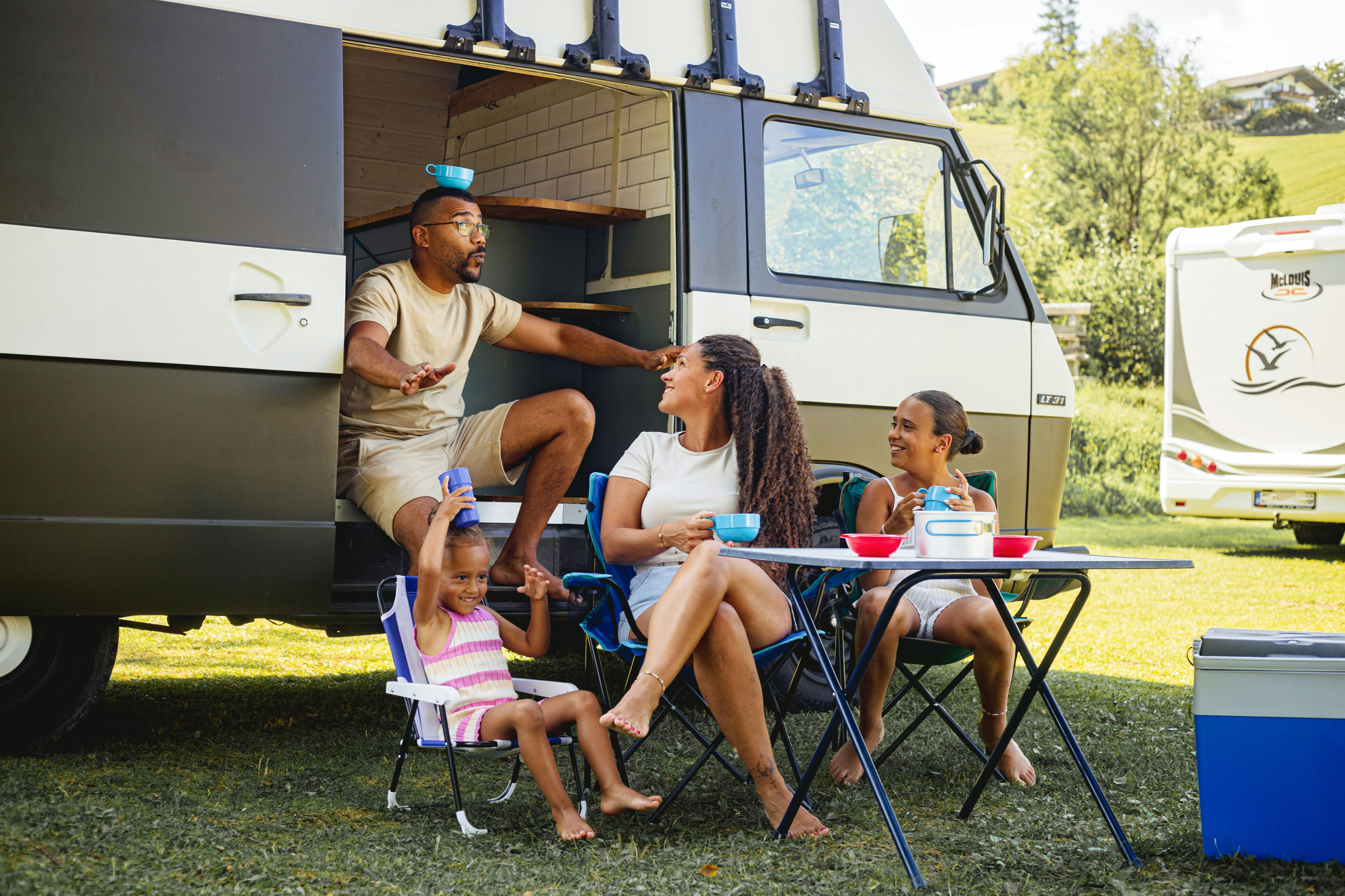 Familie sitzt am Camping-Tisch vor einem Wohnmobil, Kinder spielen mit Geschirr, im Hintergrund weitere Wohnmobile und Bäume.