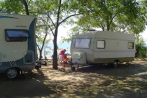 Zwei Wohnwagen auf einem Campingplatz im Schatten von Bäumen, mit Stellplatzfläche und geparkten Fahrzeugen.