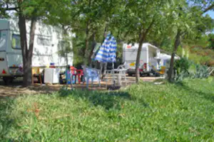 Wohnwagen und Zelt auf einem Campingplatz zwischen Bäumen, mit Stühlen und Tisch im Schatten.