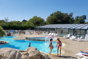 Schwimmbad mit türkisfarbenem Wasser, Sonnenschirmen und Liegen auf der Terrasse, im Hintergrund Bäume und ein Spielbereich.