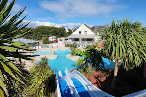 Blick auf den Camping-Pool mit Liegen, umgeben von Palmen und Pflanzen, im Hintergrund ein weißes Wohnhaus.
