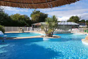 Großer Camping-Pool mit Sonnenschirmen, Liegen und Palmen; im Hintergrund ein Glasdach und Zäune.