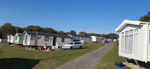 Reihen von Mobilheimen auf einem Campingplatz, Rasenfläche und Zufahrtsweg, davor ein Auto und Gartenmöbel.