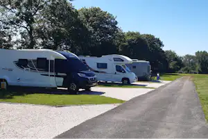 Wohnmobile und Campingfahrzeuge auf einem Stellplatz am Rand eines breiten Weges, Rasenfläche und Bäume im Hintergrund.