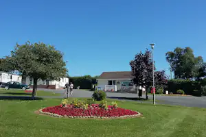 Campinganlage mit Rasen, Blumenbeet und Wegen vor einem Gebäude, daneben Bäume und geparkter Wagen.