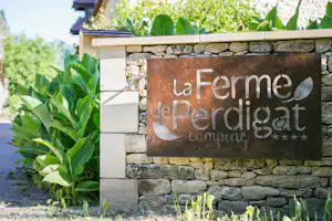 Schild „la Ferme“ am Steinmauerwerk mit grünen Pflanzen, im Hintergrund ein Gebäude und Bäume.