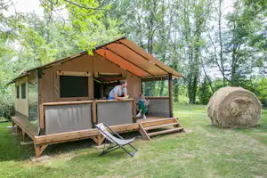 Zelt mit Terrasse auf Holzpodest im Grünen, Sonnensegel, Sitzbank und Strohballen im Garten zwischen Bäumen.