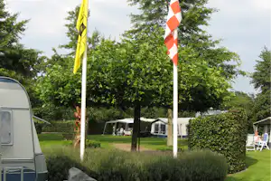 Campingplatz mit Wohnwagen am Rand, Zelte auf der Wiese und zwei Flaggen vor dichtem Grün.