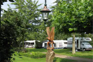 Campingplatz im Grünen: Wohnmobile und Zelte zwischen Bäumen, im Vordergrund eine Holzfigur und ein Laternenmast.