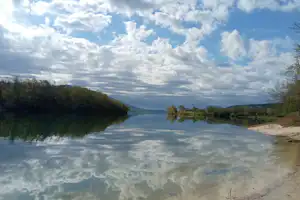 Ruhiger See mit spiegelnder Wasseroberfläche, Uferweg und Bäumen am Rand, im Hintergrund Hügel und Wolkenhimmel.