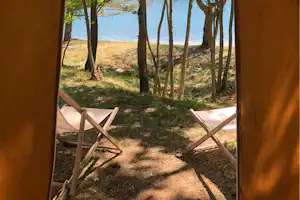 Blick aus dem Campingzelt auf den See durch Bäume, mit zwei Campingstühlen im Zeltvorraum.