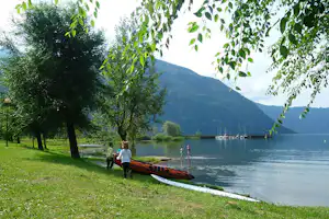 Ruderboot am Seeufer unter Bäumen, im Hintergrund Berge und mehrere Segelboote im Wasser.