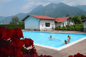 Campingpool mit blauem Wasser, daneben ein weißes Gebäude und Berge im Hintergrund, im Vordergrund rote Blumen.