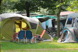 Campingzelt auf dem Stellplatz, daneben Wohnwagen und weiteres Zelt, im Hintergrund ein Pool hinter Bäumen.