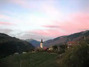 Blick ins Tal mit Kirche im Hintergrund, grünen Hängen und Bergen bei Sonnenuntergang.