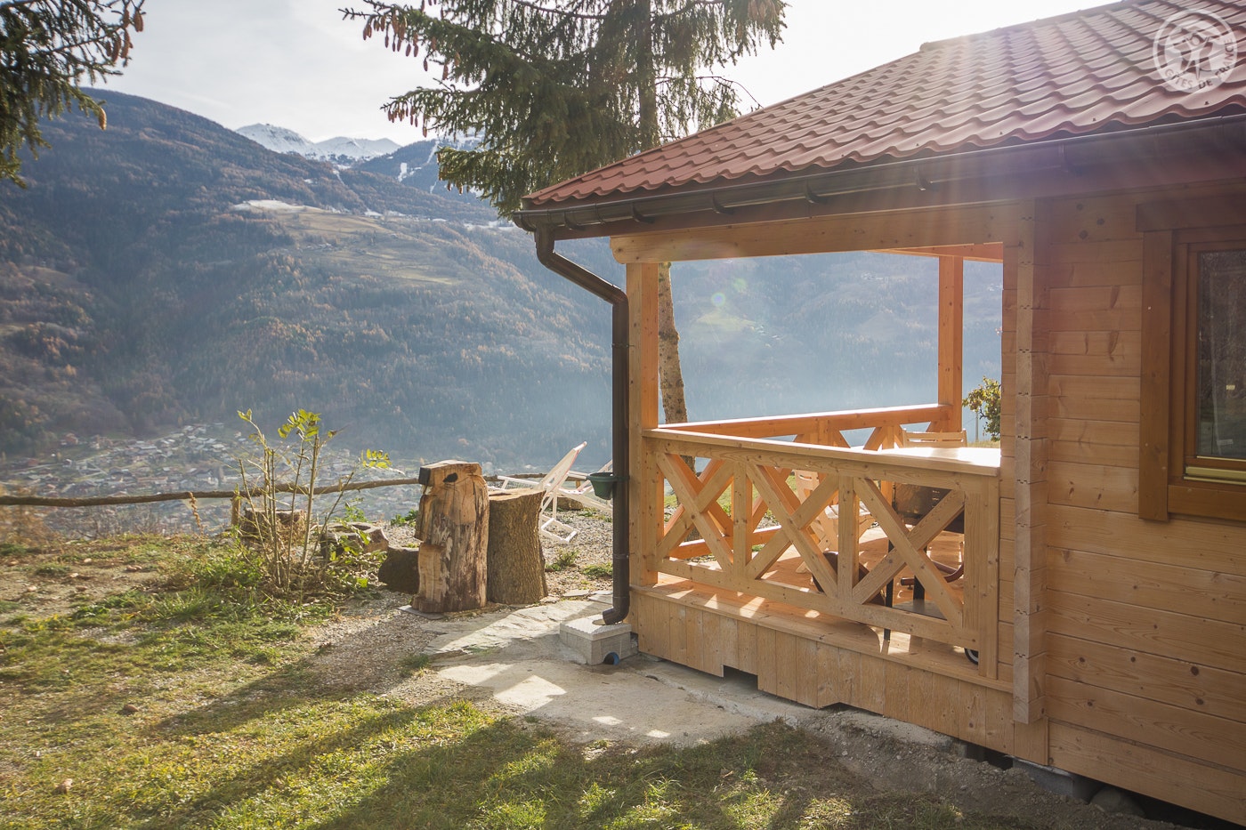 Holzhütte mit überdachter Terrasse, Geländer und Sitzbank, im Hintergrund Berge und ein Tal, am Campingplatz.