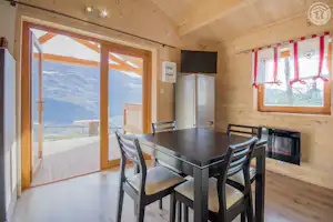 Essbereich in einer Holzhuette mit Tisch und Stuehlen, Kamin und Kuechenzeile, grosse Fenster mit Blick auf die Berge.