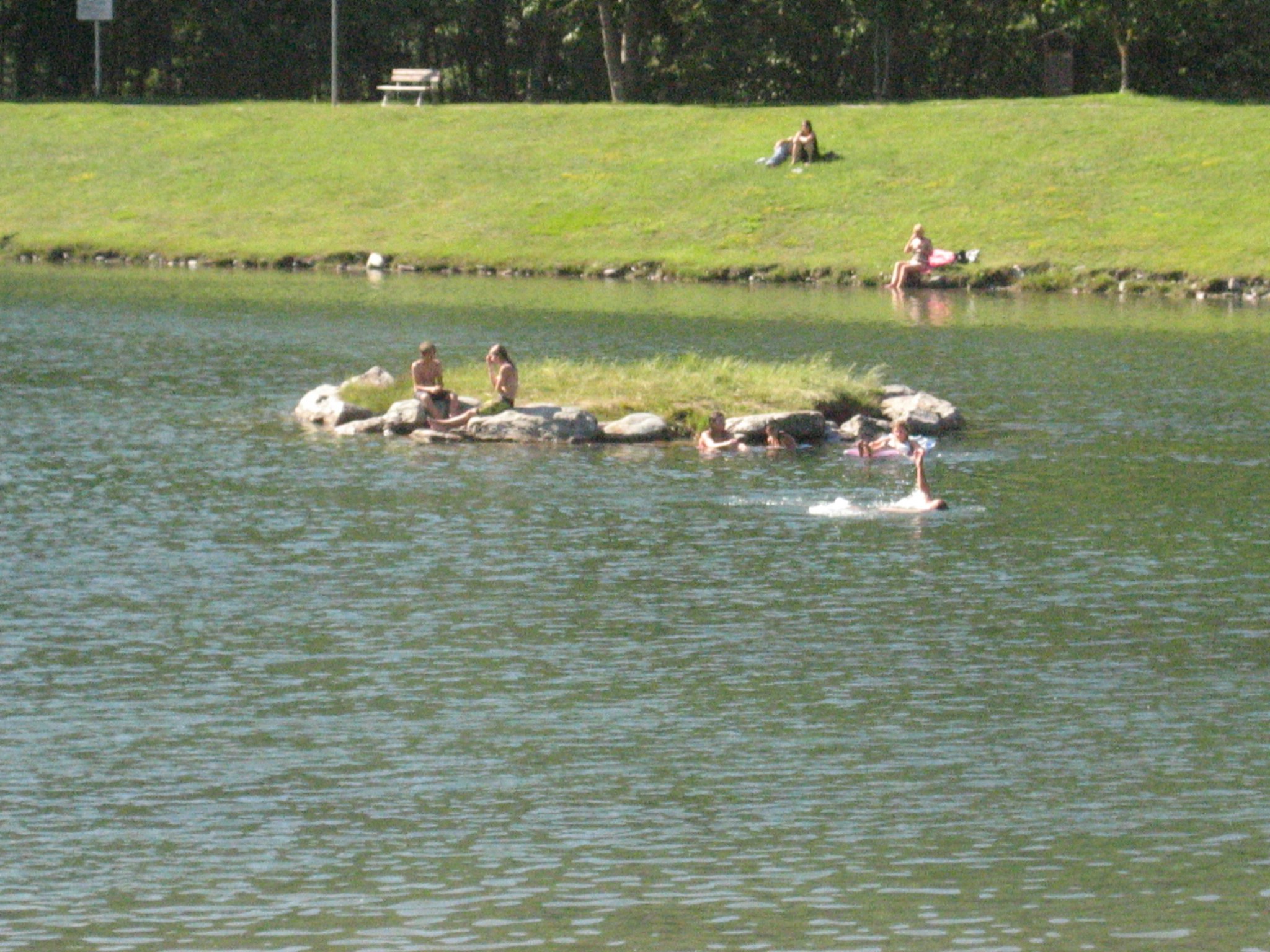 Mehrere Personen paddeln mit kleinen Booten auf einem Campingsee, im Hintergrund grünes Ufer mit Bäumen und Rasen.