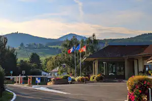 Einfahrt eines Campingplatzes mit Flaggen vor einem Gebäude, Parkplatz und Straße mit Blick auf Hügel und Bäume.