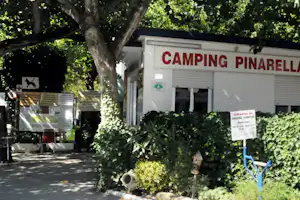 Camping-Gebäude mit rotem Schriftzug „CAMPING PINARELLA“, Rolltoren und Schildern, umgeben von Bäumen und Hecken.