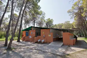 Campinggebäude mit grünem Dach und gepflastertem Vorplatz im Wald, neben Mülltonnen und Schildern.