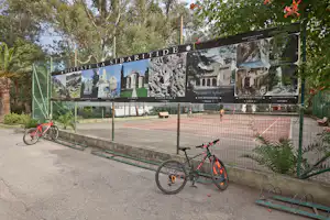 Fahrrad vor einer großen Infotafel am Campingplatz, im Hintergrund Bäume und ein umzäunter Bereich mit Bänken.