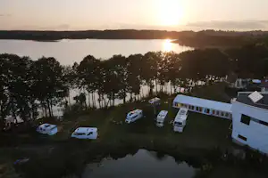 Campingplatz am See mit Wohnwagen und Stellplätzen am Ufer, im Hintergrund Bäume und die untergehende Sonne.