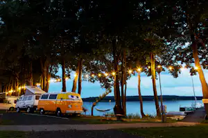 Wohnwagen und gelber Camper am Seeufer im Abendlicht, Bäume, Holzweg und Bootssteg mit Booten.