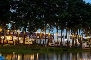 Campingplatz am See mit beleuchteten Stellplätzen, Wohnwagen und Bäumen am Ufer bei Abendlicht.