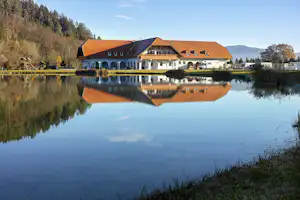 Campinghaus am See mit Spiegelung im Wasser, Satteldach und Arkaden, im Hintergrund Wald und Berge.