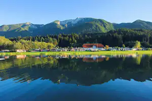 Campingplatz am See mit ruhigem Wasser, Bootssteg und Wohnwagen, im Hintergrund Wald und Berge.