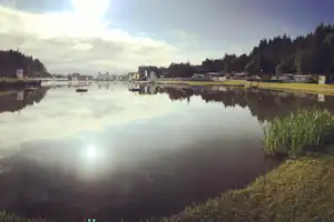 Ruhiger Campingsee mit Spiegelung von Gebäuden und Booten, im Hintergrund Wald und Ufergras.