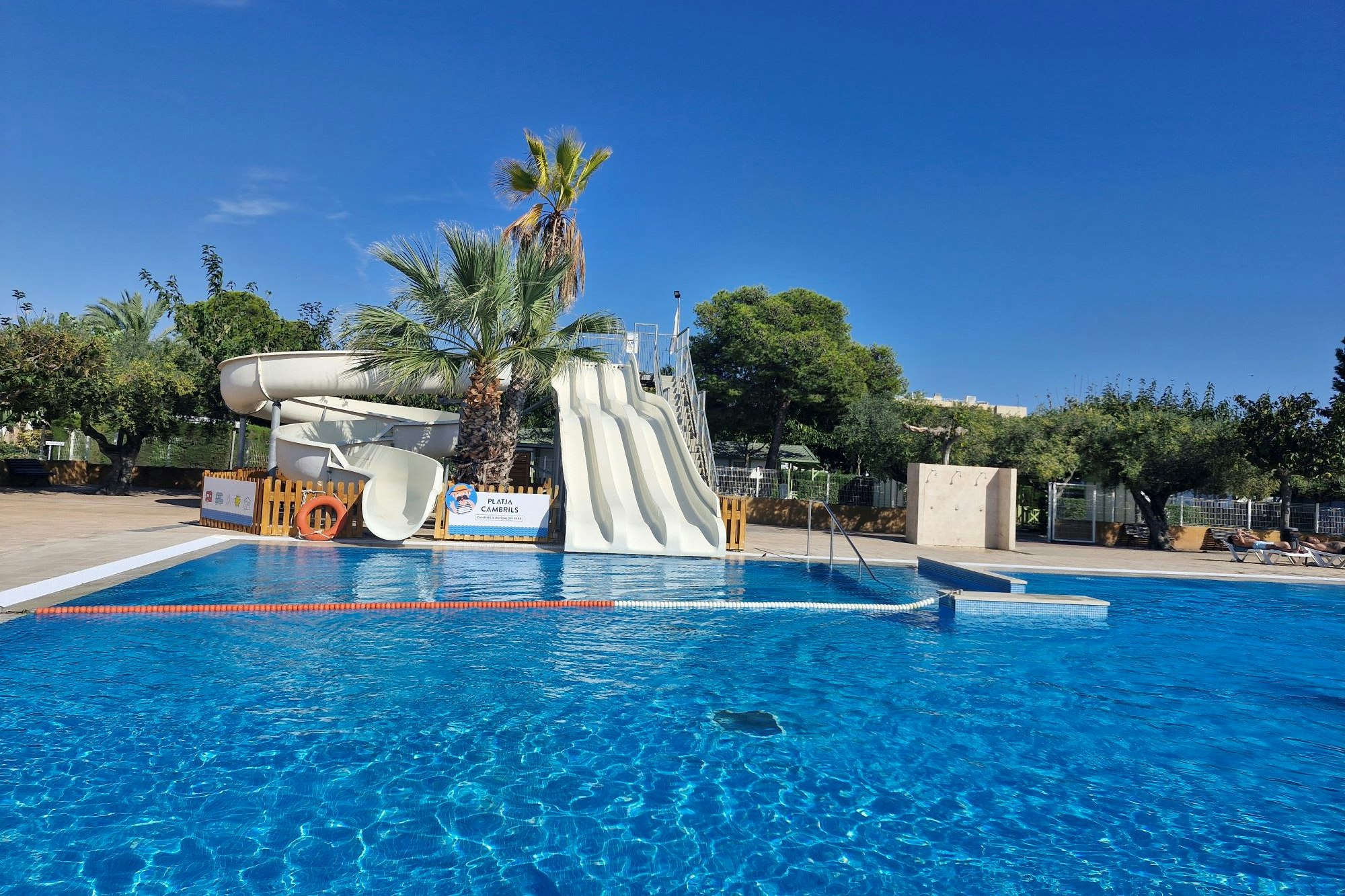 Blaues Schwimmbecken mit Liegen und Sonnenschirm, im Hintergrund ein weißes Wasserrutschen- und Spielbereich am Campingplatz.