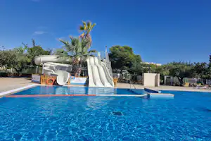 Blaues Schwimmbecken mit Liegen und Sonnenschirm, im Hintergrund ein weißes Wasserrutschen- und Spielbereich am Campingplatz.
