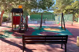 Spielplatz im Campingbereich mit Schaukel, Klettergerüst und buntem Fallschutzboden, umgeben von hohen Bäumen und Zäunen.