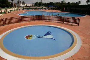 Rundes Camping-Poolbecken mit Delfin-Mosaik, umgeben von Holzzaun und Terrassenfliesen, im Hintergrund ein weiteres Schwimmbecken.