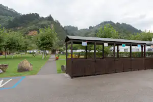 Campinganlage mit Rasen, Baumreihen und gepflastertem Weg, im Hintergrund Berge und ein Holzunterstand mit Schildern.