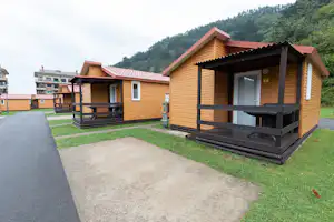 Holzbungalow mit Terrasse, Eingangstür und Fenstern, davor Holzgeländer und Bank, gepflasterter Weg und Rasen am Campingplatz.
