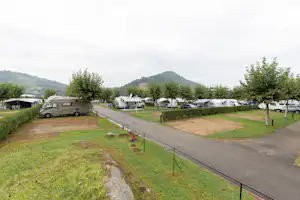 Wohnmobile und Campingbusse auf einem Campingplatz, gepflasterter Weg, Rasenflächen und Hecken, im Hintergrund grüne Hügel.