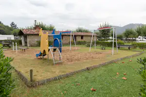 Spielplatz im Campinggarten: bunte Kletterwand und Schaukel auf Sand, Bänke und Hecken neben einem Steingebäude.
