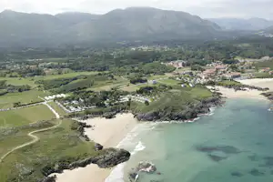 Luftaufnahme einer Küstenbucht mit Sandstrand, grünen Hängen, Häusern und Bergen im Hintergrund.