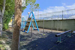 Spielplatz auf dem Campingplatz mit blauer Rutsche, Zaun und gepflastertem Weg zwischen Bäumen.