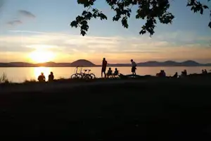 Sonnenuntergang am Seeufer: Menschen sitzen und stehen am Steg, im Hintergrund Berge und Wasser.