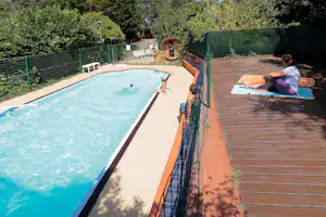 Camping-Pool mit blauem Wasser, Holzdeck und Liegen, im Hintergrund Hecken, Zaun und ein kleines Holzhaus.