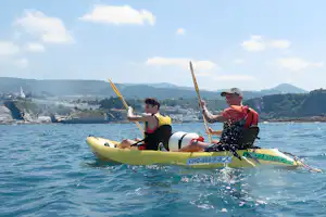 Zwei Personen im Kajak mit Paddeln auf dem See, im Hintergrund Küste, Häuser und bewaldete Hügel.
