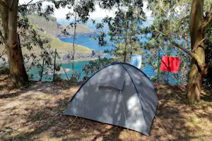 Zelt im Schatten unter Bäumen am Campingplatz mit Blick auf das Meer und die Küste in den Bergen.