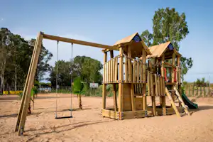 Holz-Spielplatz mit Klettergerüst und Schaukeln auf Sandboden, im Hintergrund Bäume und Zaun.