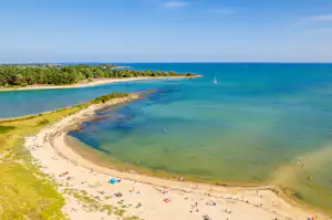 Luftaufnahme einer Sandbucht mit klarem Meer, Küstenlinie, Bäumen und vielen Strandbesuchern am Ufer.