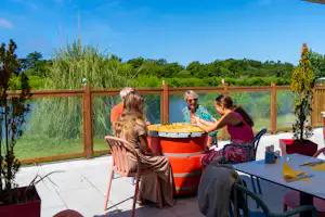 Terrasse am Campingplatz mit Whirlpool, Holzzaun und Blick auf den See, im Hintergrund Bäume und grünes Ufer.