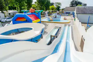Familienpool mit blauen Rutschen, Planschbecken und Spielbereich, umgeben von Terrasse, Bäumen und Wohngebäuden.